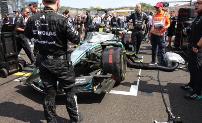 Mercedes race car in pit lane with crew members and engineers working on it, banners and grandstands in the background