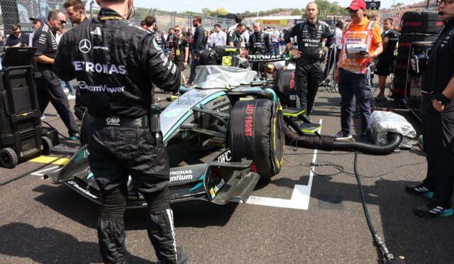 Mercedes race car in pit lane with crew members and engineers working on it, banners and grandstands in the background
