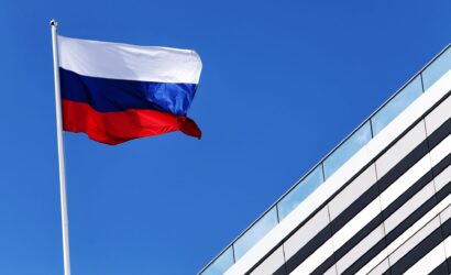 Russian national flag fluttering on a flagpole against a clear blue sky beside a modern glass office building.