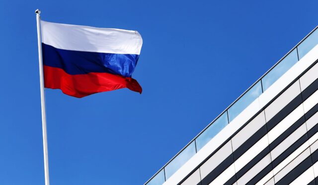 Russian national flag fluttering on a flagpole against a clear blue sky beside a modern glass office building.