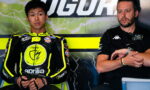 MotoGP rider in yellow-black Aprilia suit sits in the pit area beside a team member in a black polo.