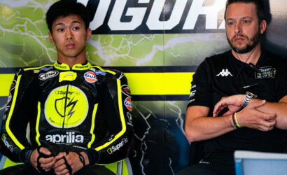 MotoGP rider in yellow-black Aprilia suit sits in the pit area beside a team member in a black polo.