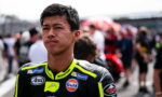 Young male motorcycle racer in neon-yellow and black suit, standing on a track with a blurred crowd in the background.