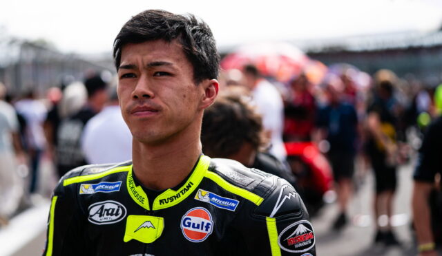 Young male motorcycle racer in neon-yellow and black suit, standing on a track with a blurred crowd in the background.