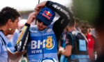 Moto racer in blue sponsor jersey wearing Red Bull cap adjusts helmet with help from team member amid a crowded pit area