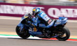 MotoGP rider on a blue racing bike leaning into a turn on a race track, sponsor livery visible across suit and bike.