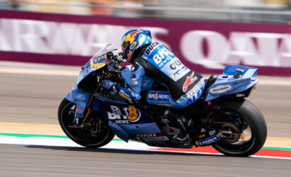 MotoGP rider on a blue racing bike leaning into a turn on a race track, sponsor livery visible across suit and bike.
