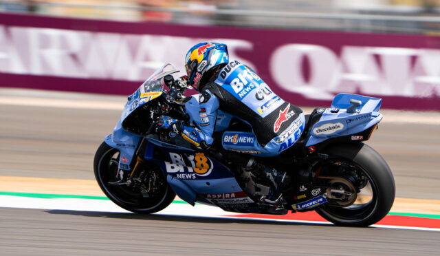 MotoGP rider on a blue racing bike leaning into a turn on a race track, sponsor livery visible across suit and bike.