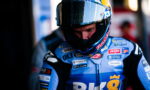 Motorcycle racer in a blue sponsor-painted racing suit and full-face helmet, looking down in the pit area with logos on the suit.