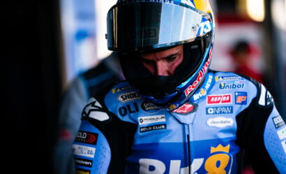 Motorcycle racer in a blue sponsor-painted racing suit and full-face helmet, looking down in the pit area with logos on the suit.