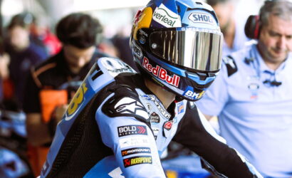 MotoGP rider in a blue racing suit wearing a helmet with Red Bull and Shoei logos, in the pit area.