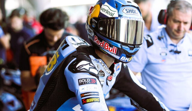 MotoGP rider in a blue racing suit wearing a helmet with Red Bull and Shoei logos, in the pit area.