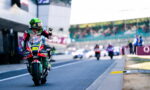 Motocycle racer wearing a bright green helmet on a red-green racing bike, giving a thumbs-up on a race track with blurred supporters in the background.