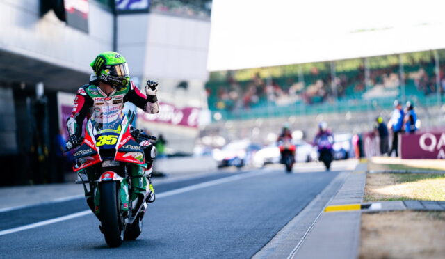 Motocycle racer wearing a bright green helmet on a red-green racing bike, giving a thumbs-up on a race track with blurred supporters in the background.