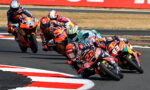 Group of motorcycle racers leaning into a sharp turn on a race track, colorful sponsor livery visible on bikes.