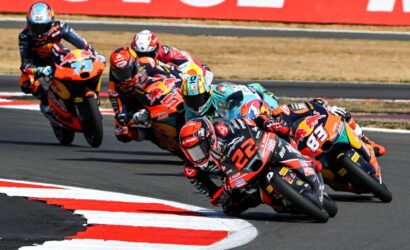 Group of motorcycle racers leaning into a sharp turn on a race track, colorful sponsor livery visible on bikes.