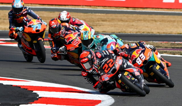 Group of motorcycle racers leaning into a sharp turn on a race track, colorful sponsor livery visible on bikes.