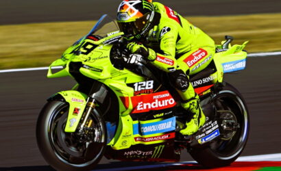 Racing motorbike in neon yellow livery leaning into a track bend with a helmeted rider in matching gear, sponsors visible on bike sides and fairings.