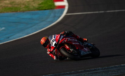 Red motorcycle racer in red suit leans deeply into a left-hand turn on a dark race track with blue runoff area nearby.