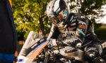 Racer in full suit and helmet hunched over a Ducati race bike, sponsor stickers visible, on a sunlit pit area backdrop.