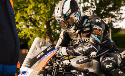 Racer in full suit and helmet hunched over a Ducati race bike, sponsor stickers visible, on a sunlit pit area backdrop.