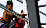 Motocross rider shirtless on scaffolding, wearing a Monster cap and sunglasses, holding a water bottle.