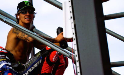 Motocross rider shirtless on scaffolding, wearing a Monster cap and sunglasses, holding a water bottle.