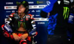 MotoGP rider in Yamaha gear sits in the pit area wearing a headset, looking to the side.