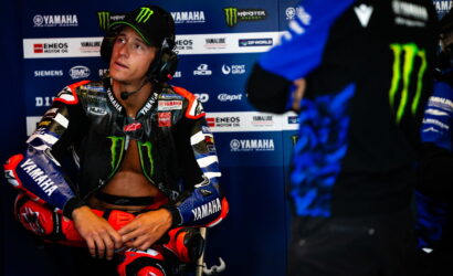 MotoGP rider in Yamaha gear sits in the pit area wearing a headset, looking to the side.