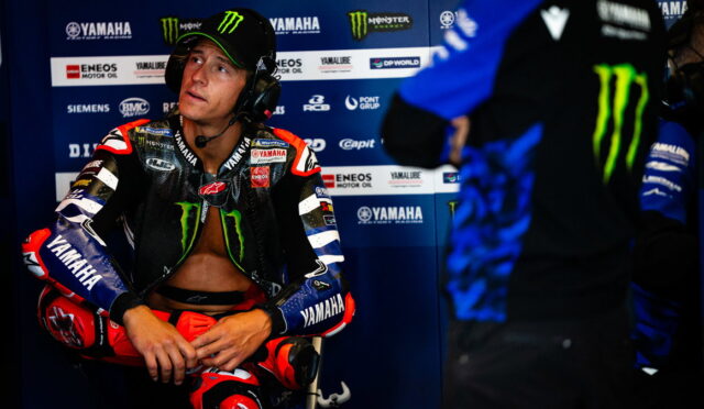 MotoGP rider in Yamaha gear sits in the pit area wearing a headset, looking to the side.