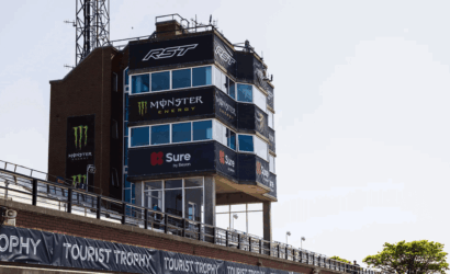Racing control tower at a track covered with sponsor banners (Monster Energy, Sure) and Tourist Trophy signage along the wall.