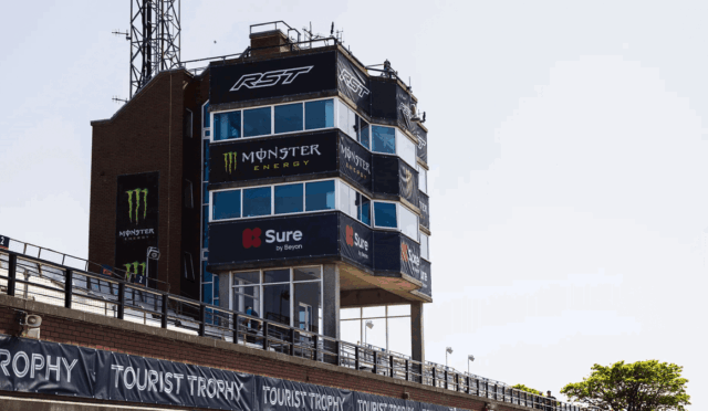 Racing control tower at a track covered with sponsor banners (Monster Energy, Sure) and Tourist Trophy signage along the wall.