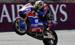 Motorcycle racer in colorful livery performing a wheelie on a racing bike during a track event, sponsor logos visible on the bike ground in the background
