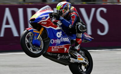 Motorcycle racer in colorful livery performing a wheelie on a racing bike during a track event, sponsor logos visible on the bike ground in the background