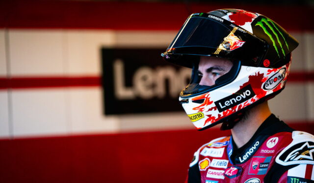 Racer wearing a full-face helmet with Lenovo logo in a garage, neon red and white background behind him.