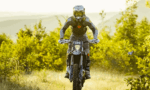 Off-road motorcycle rider in full gear performing a jump toward the camera on a dirt trail, surrounded by foliage.",