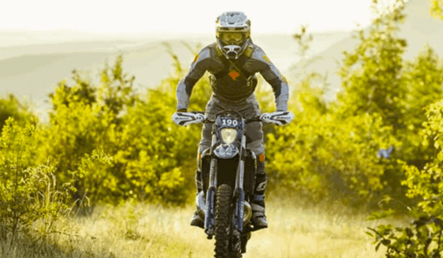 Off-road motorcycle rider in full gear performing a jump toward the camera on a dirt trail, surrounded by foliage.",