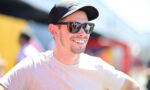 Man wearing a baseball cap and sunglasses, smiling outdoors on a sunny day.