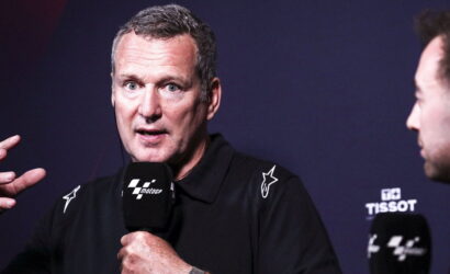 Man in a black polo speaks into a MotoGP microphone during a press event, left hand raised for emphasis.