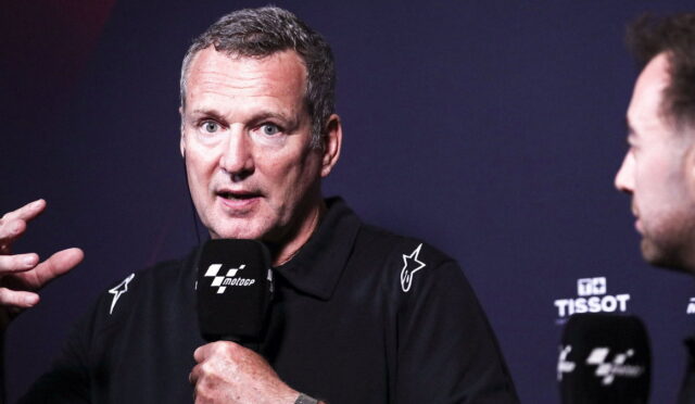 Man in a black polo speaks into a MotoGP microphone during a press event, left hand raised for emphasis.