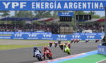 Motorcycle race on a track with riders in colorful leathers, blue YPF banners above and along the sidelines in Argentina.