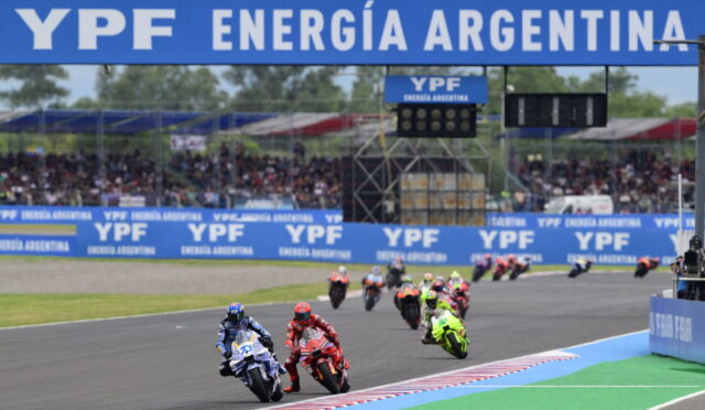 Motorcycle race on a track with riders in colorful leathers, blue YPF banners above and along the sidelines in Argentina.