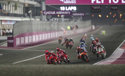 Motorcycle race on a curving track with riders leaning into a turn; Qatar Airways banners overhead.