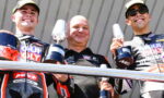 Three motorbike racers on a podium smiling, each holding a trophy in their hands.