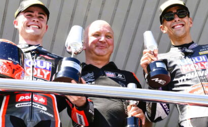 Three motorbike racers on a podium smiling, each holding a trophy in their hands.