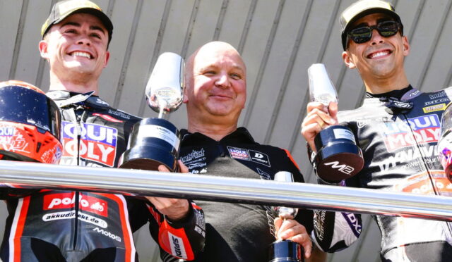 Three motorbike racers on a podium smiling, each holding a trophy in their hands.