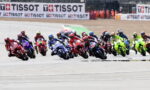 MotoGP-style pack of riders leaning into a turn on a race track, colorful bikes and TISSOT banners in the background