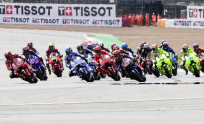 MotoGP-style pack of riders leaning into a turn on a race track, colorful bikes and TISSOT banners in the background