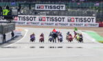 Motorcycle racers line up at the track start with a marshal raising red and green flags, behind large TISSOT banners and spectators in the stands.