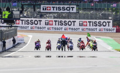 Motorcycle racers line up at the track start with a marshal raising red and green flags, behind large TISSOT banners and spectators in the stands.